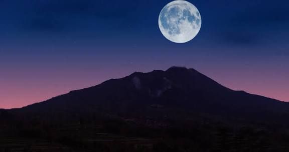 Volle maan boven een donker bergsilhouet in de schemering met een paarsroze hemel.