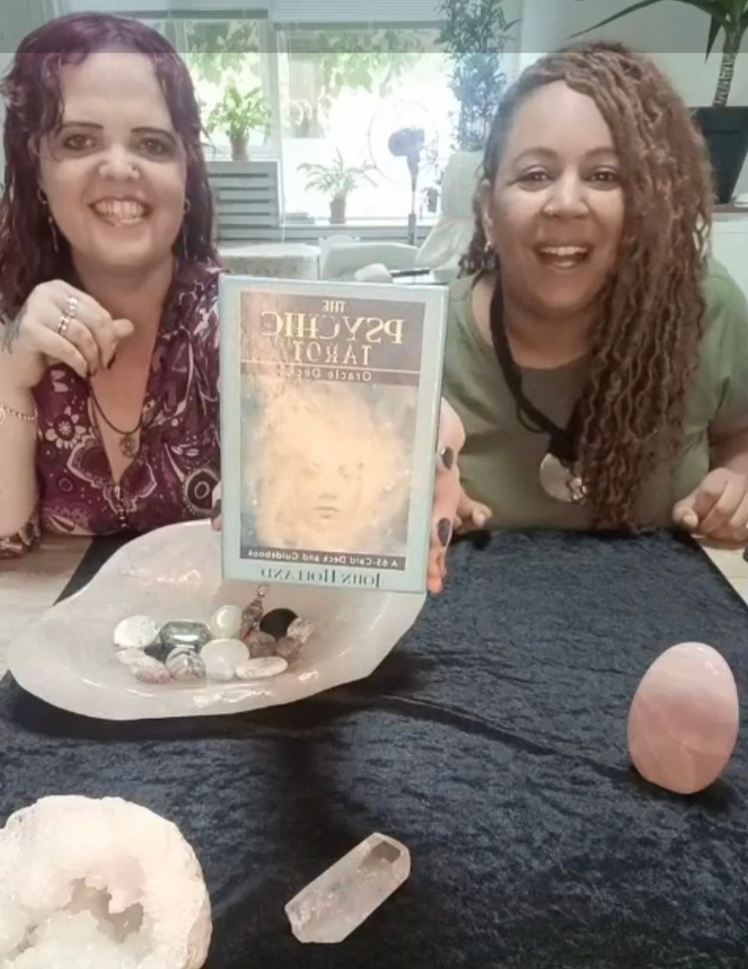 Twee lachende vrouwen zitten aan een tafel met kristallen en een display met tarot-/orakelkaarten.