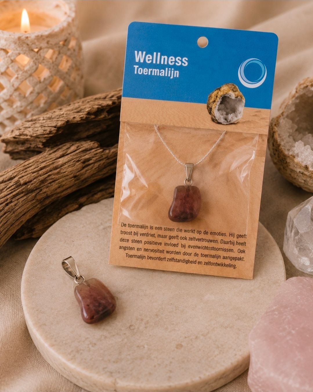 Een wellnessketting op een beige presentatiekaartje, naast een bijpassende hanger en kaars op een zachte stenen ondergrond.
