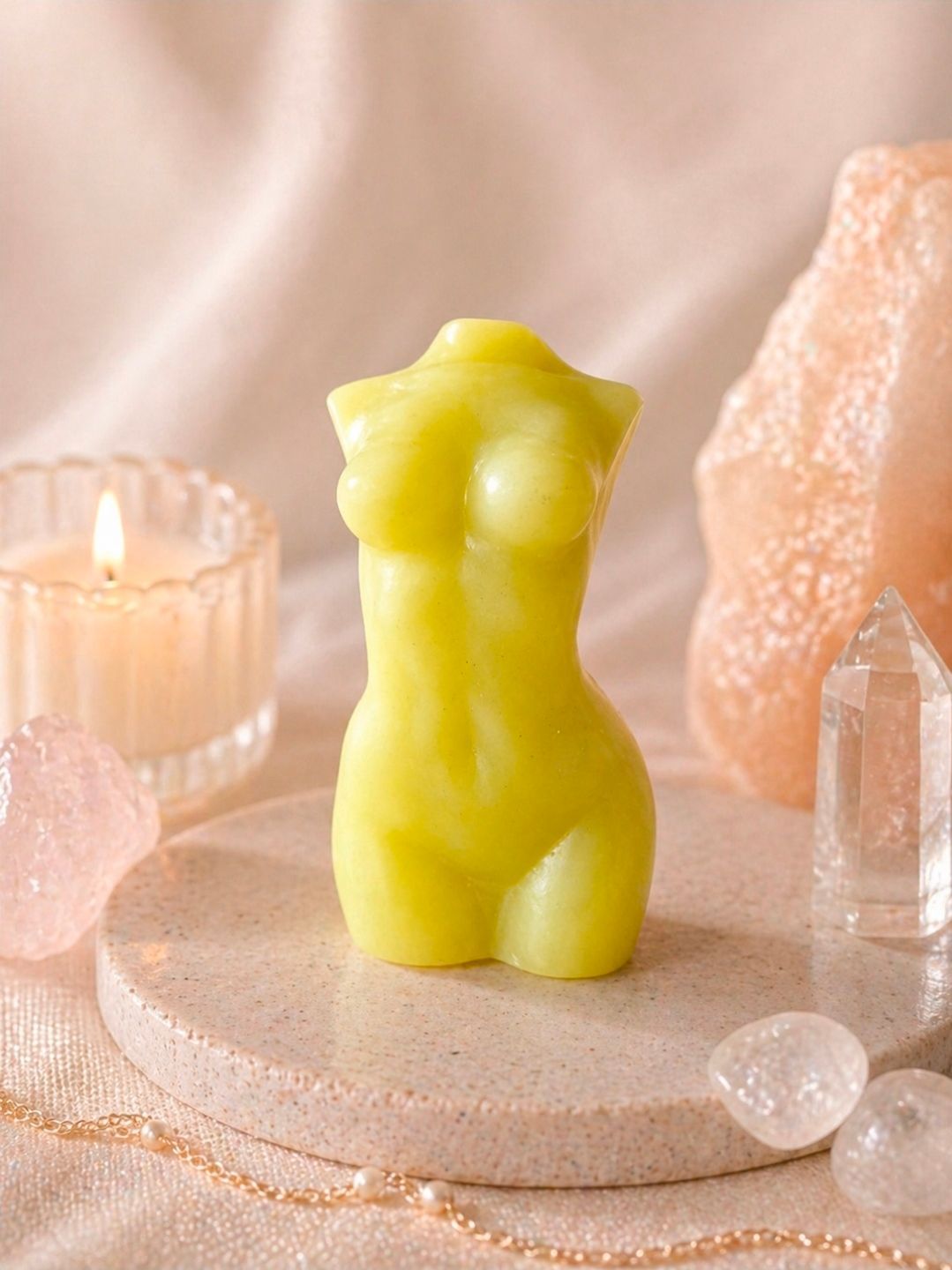Een gele kaars in de vorm van een torso op een roze stenen schaal met kristallen en een brandende kaars in zachtroze licht.