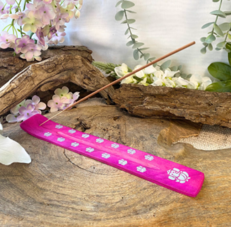 Roze wierookhouder met een brandend wierookstokje op een houten ondergrond, met bloemen en groen op de achtergrond.