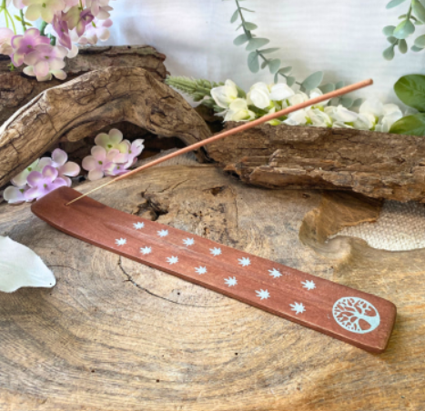 Wierookstokje in een houten houder op drijfhout, met bloemen en een kleine hagedis in een natuurlijke setting.