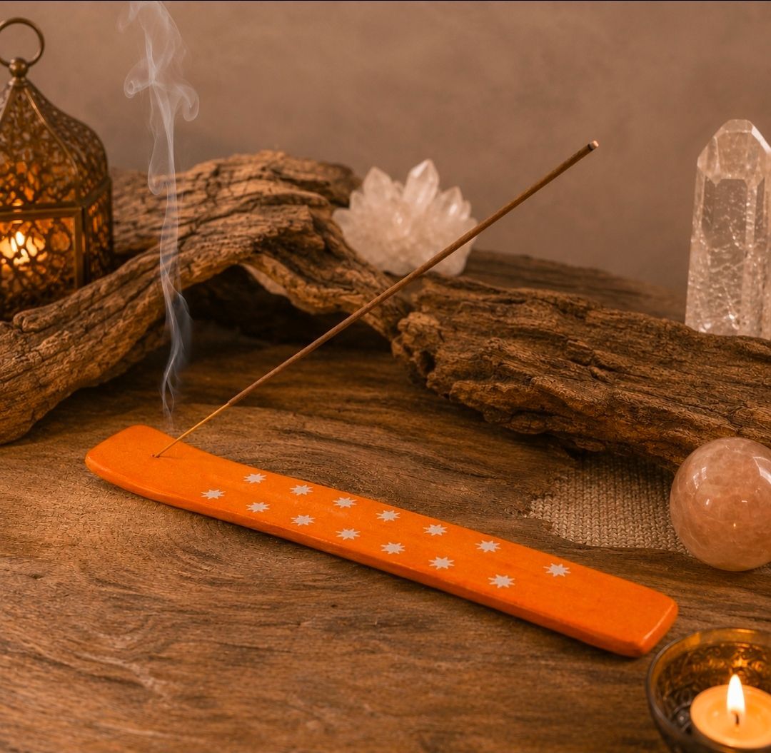 Een oranje wierookstokje brandt op een houten ondergrond met kristallen, drijfhout en een kaars in een warme omgeving.