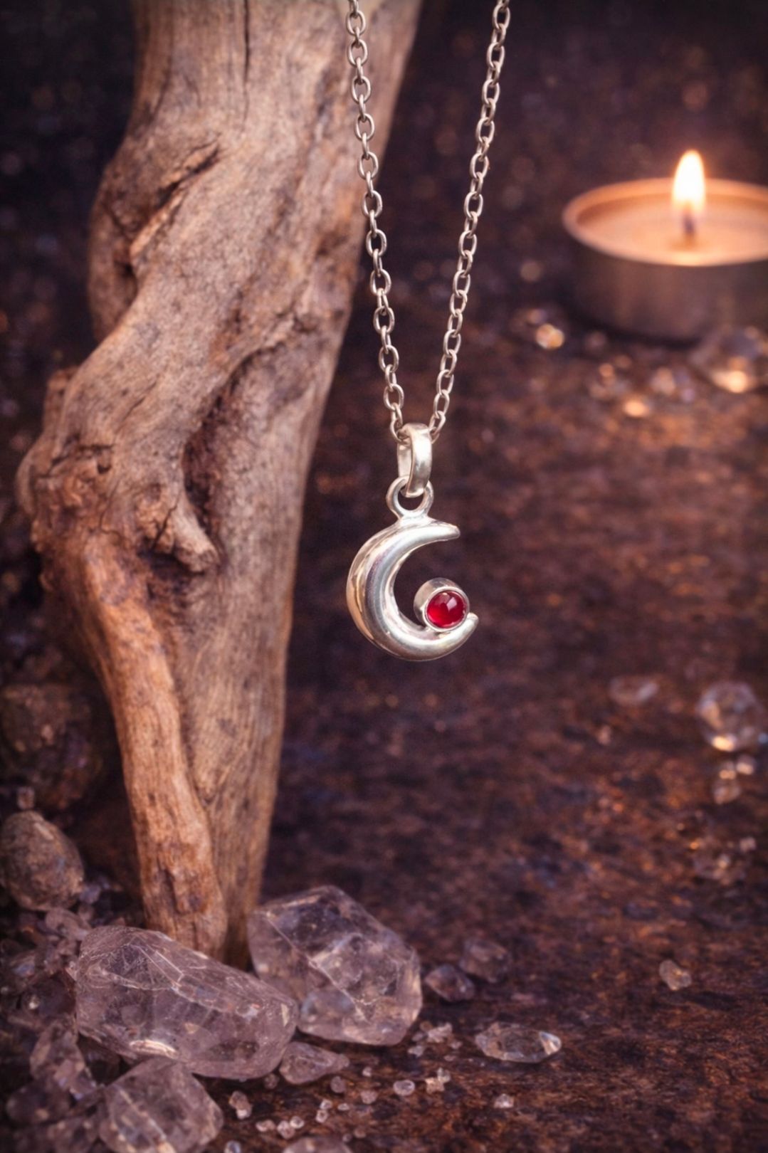 Zilveren halvemaanvormige hanger met rode edelsteen aan een ketting, naast hout en kaarslicht.