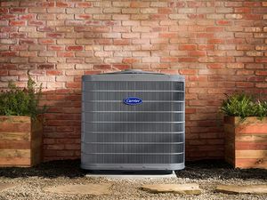 Outdoor air conditioner unit against a brick wall, flanked by potted plants on a gravel bed.