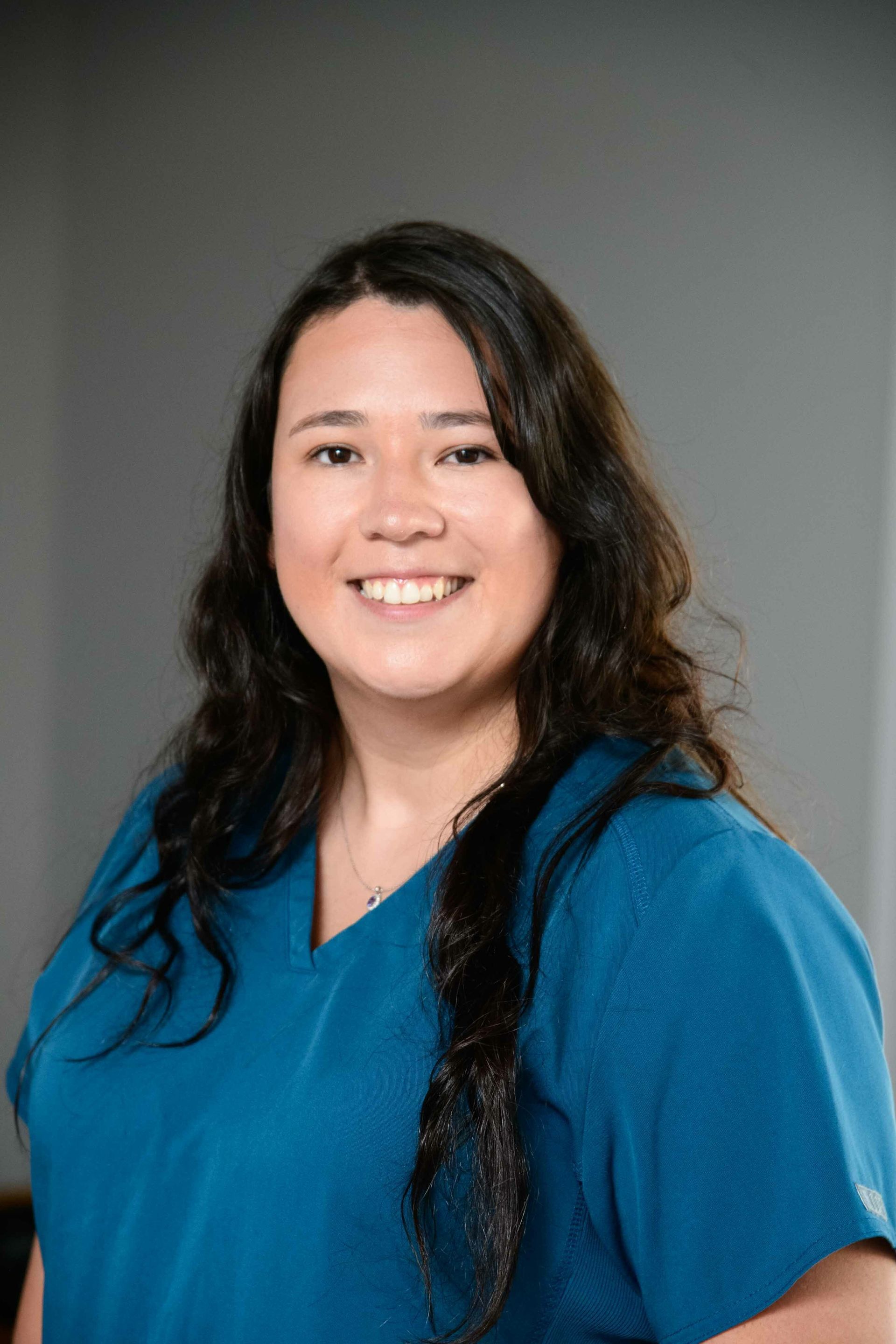 Woman with long dark hair, smiling, wearing a blue v-neck top. Neutral background.