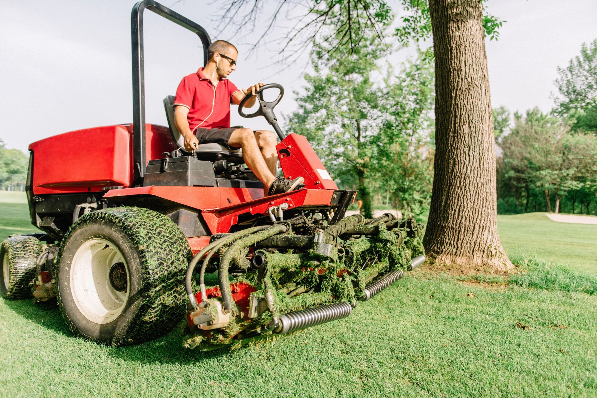Man in red shirt operates a red riding lawnmower, trimming grass near a tree in a sunny park.