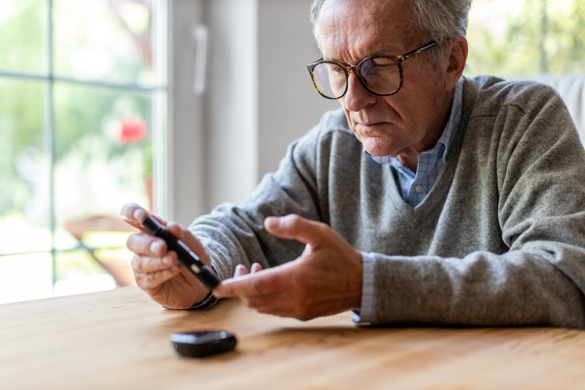 Older man wearing glasses and a gray sweater sits at a tableusing a blood glucose meter to check his blood sugar level.