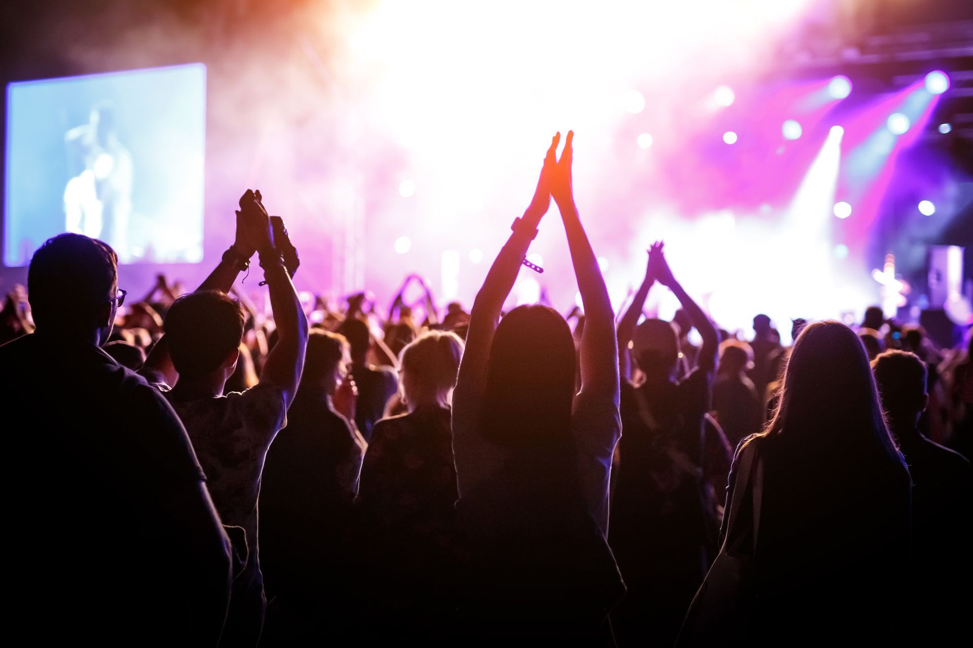 People with raised hands cheering at a brightly lit concert stage.