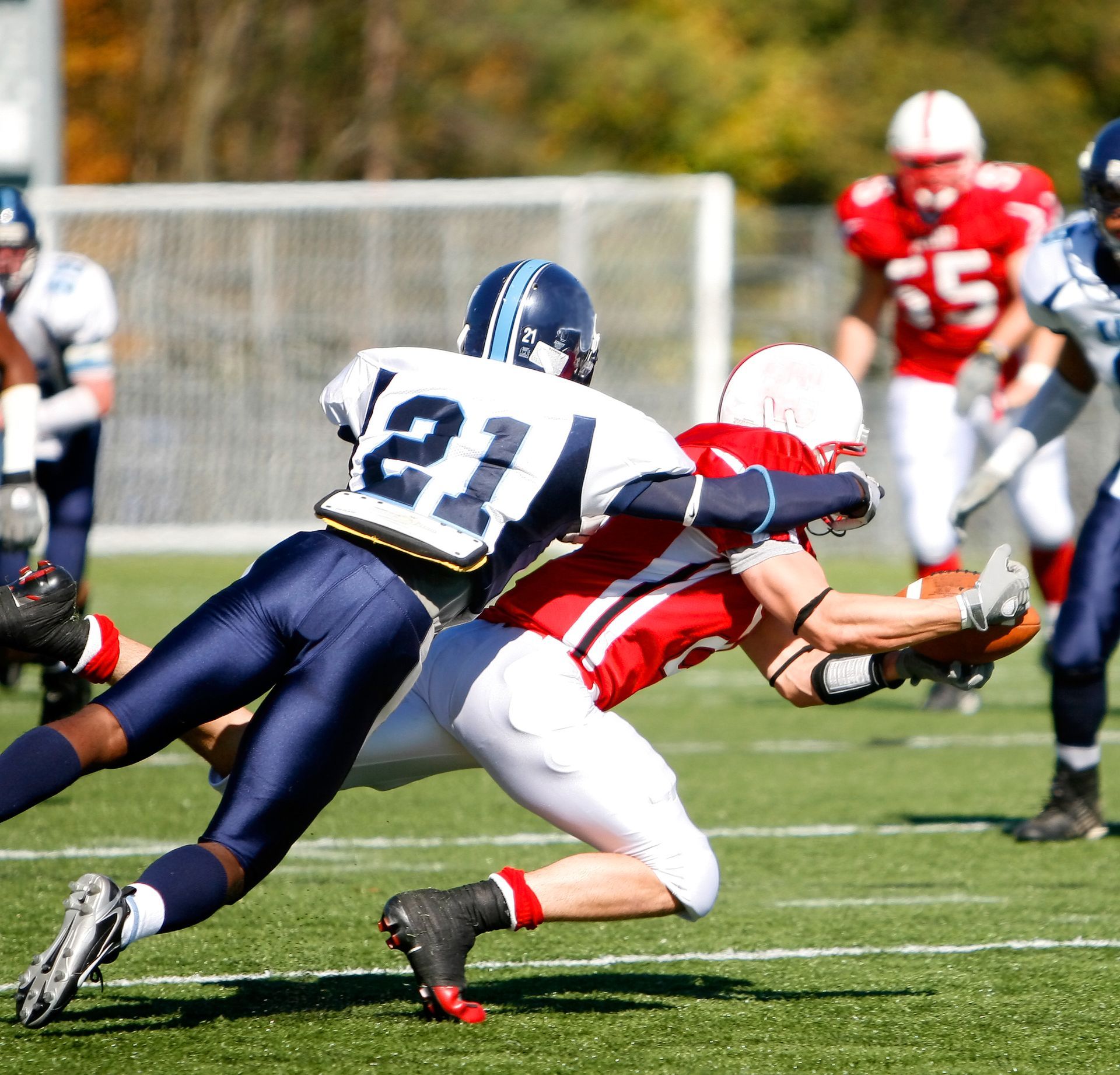 Football players in action on a green field. One is tackling another while holding the ball, others watch.