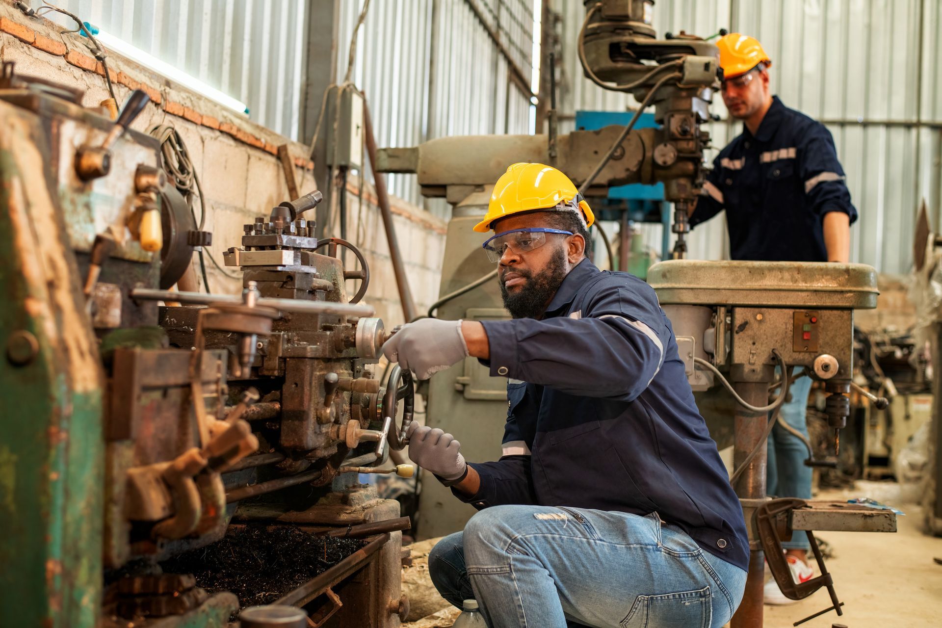 Two men in a factory working on machinery, wearing hard hats. One kneels while operating equipment.