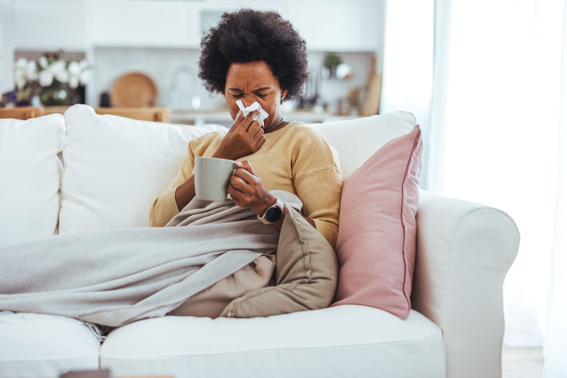 Woman with a cold, blowing nose and drinking tea on a couch.