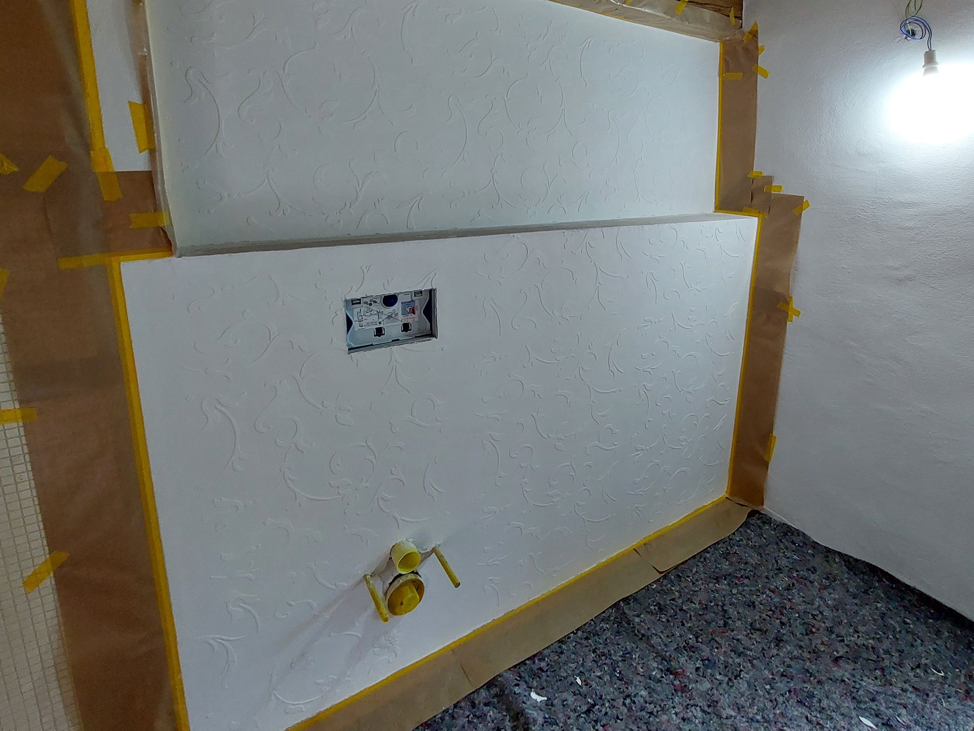 Una stanza con un muro bianco, un vano tecnico sanitario e un nastro giallo sul pavimento.