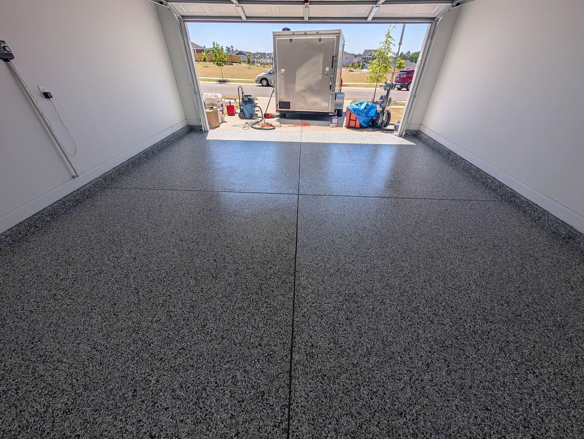 Empty garage with speckled epoxy floor and open door to a driveway with cleaning supplies outside