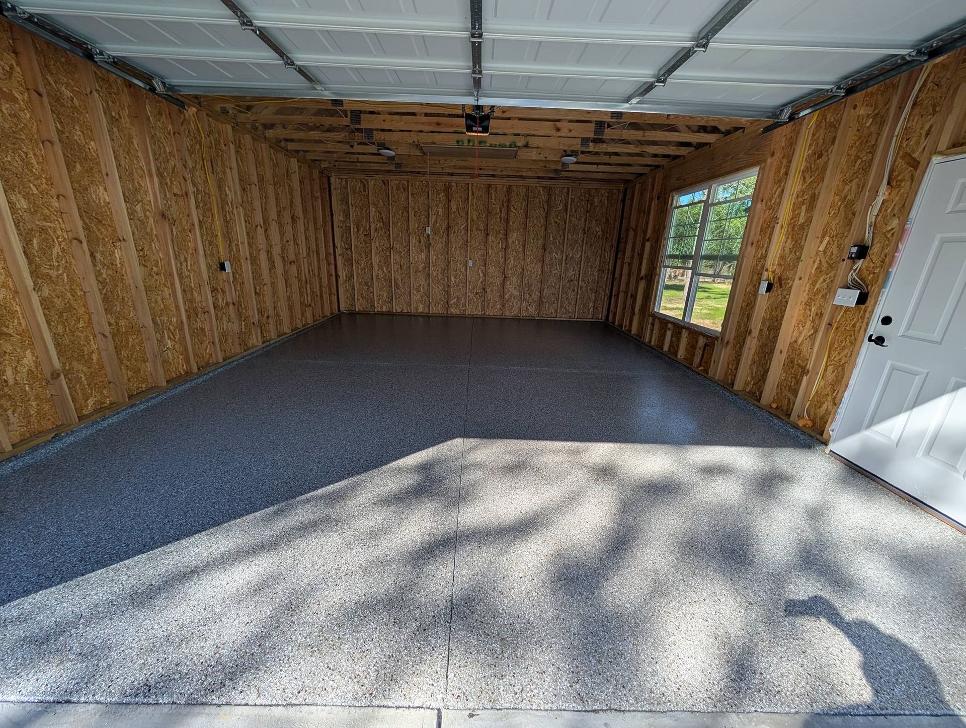 Empty unfinished garage with plywood walls, gray carpet floor, and sunlight through the open door.