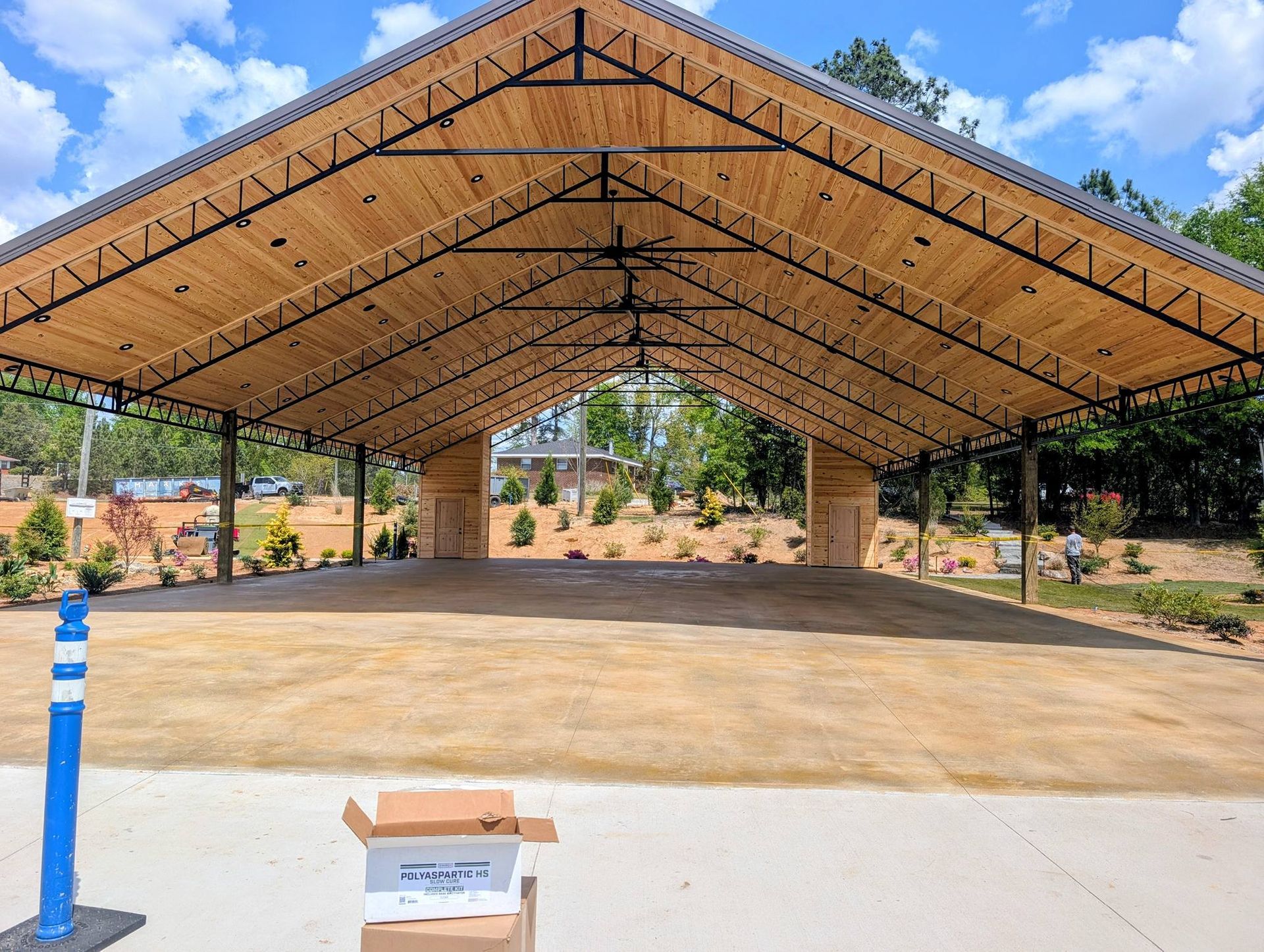 Covered open-air pavilion with wooden roof, concrete floor, and blue bollard in front under a clear sky