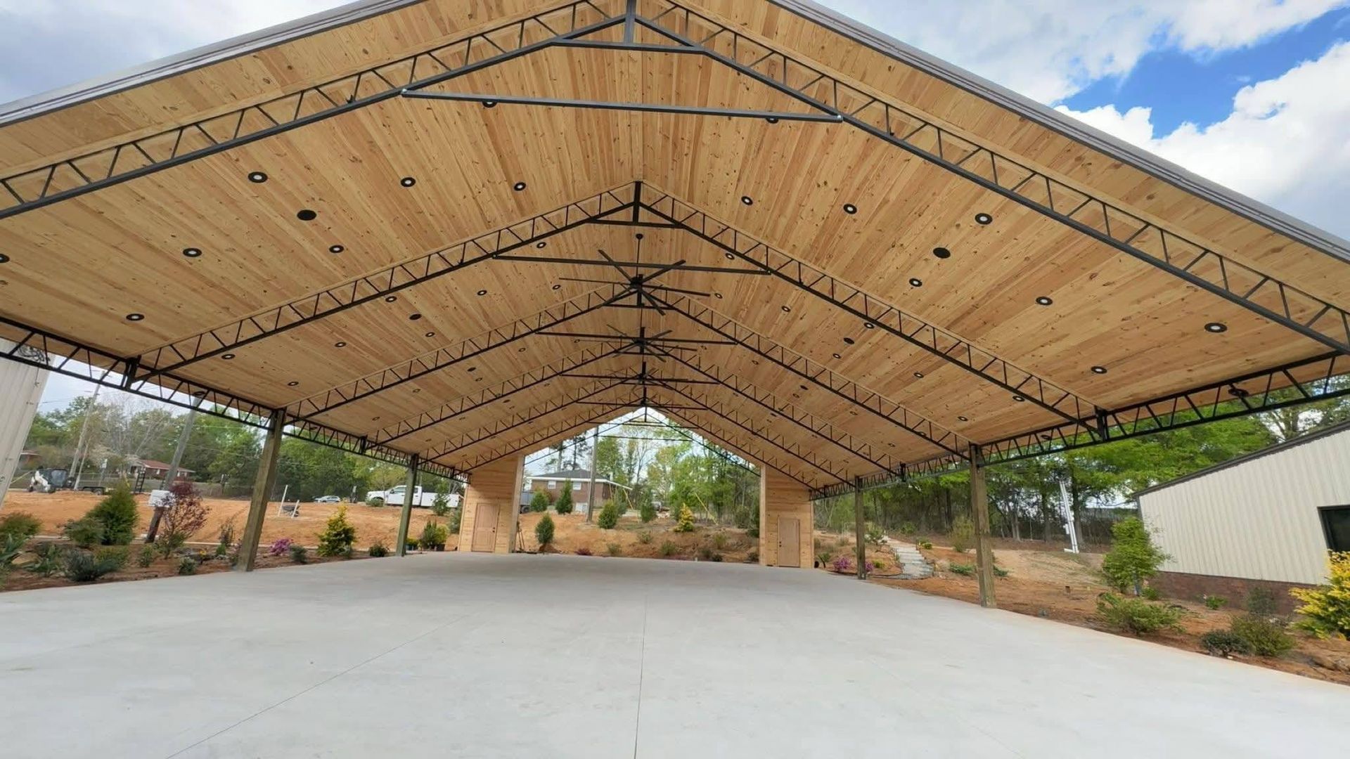 Large open-air pavilion with a high wooden truss roof over a concrete floor, surrounded by landscaped greenery.