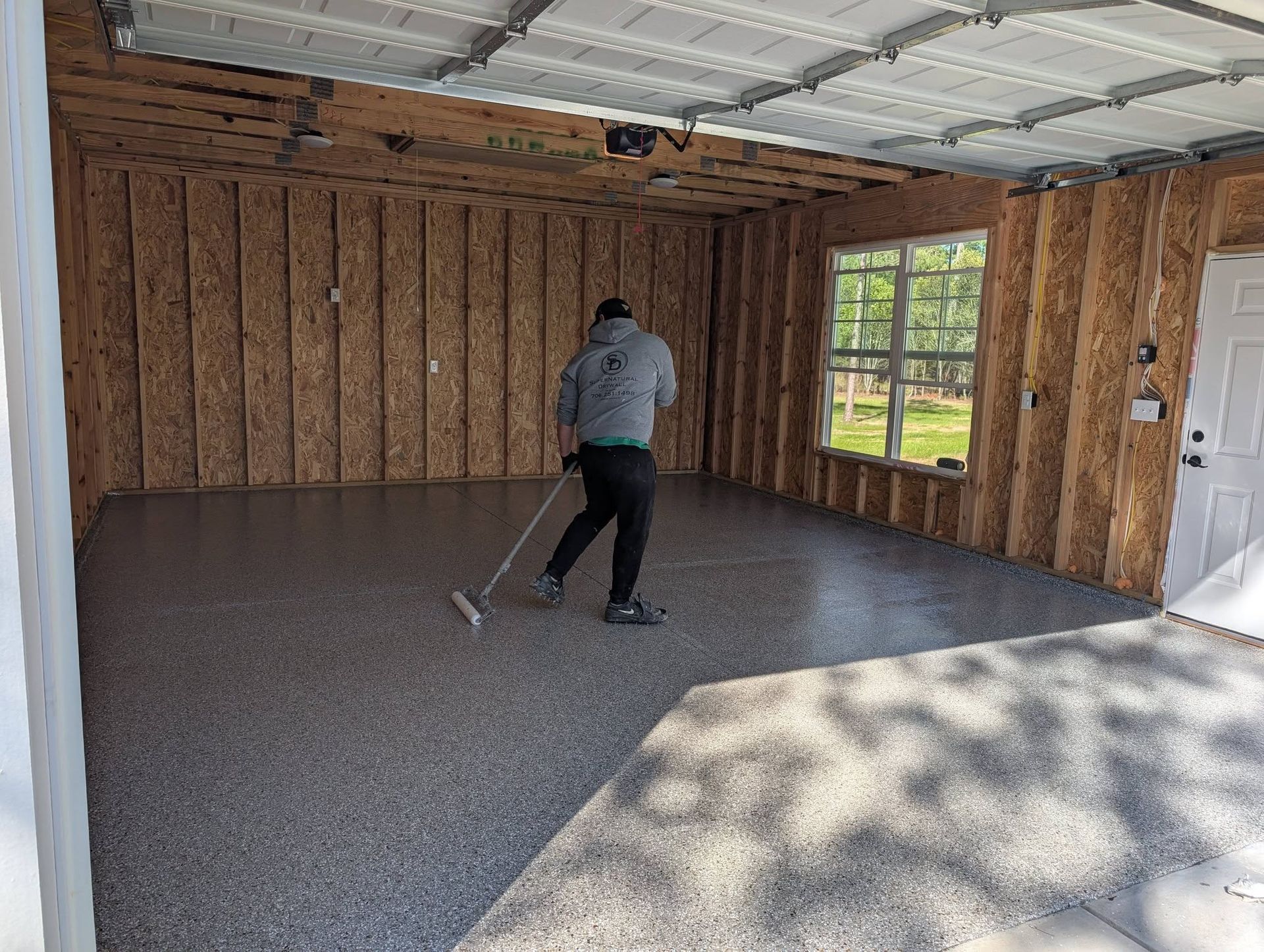Person using a push broom to sweep a bare garage with exposed studs and sunlight through a window.