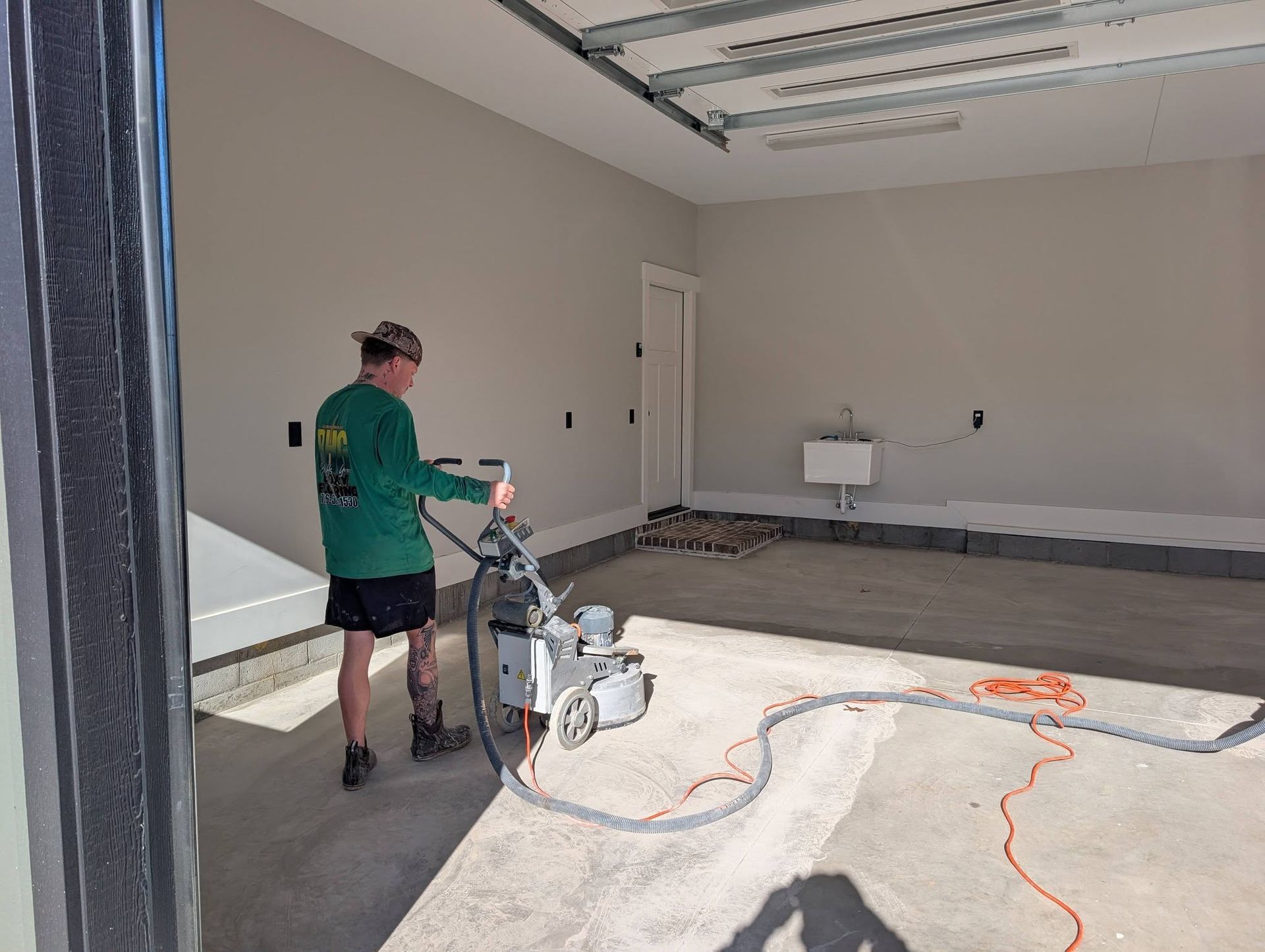Worker using a floor grinder in a bare garage under renovation, with extension cords on the concrete floor.