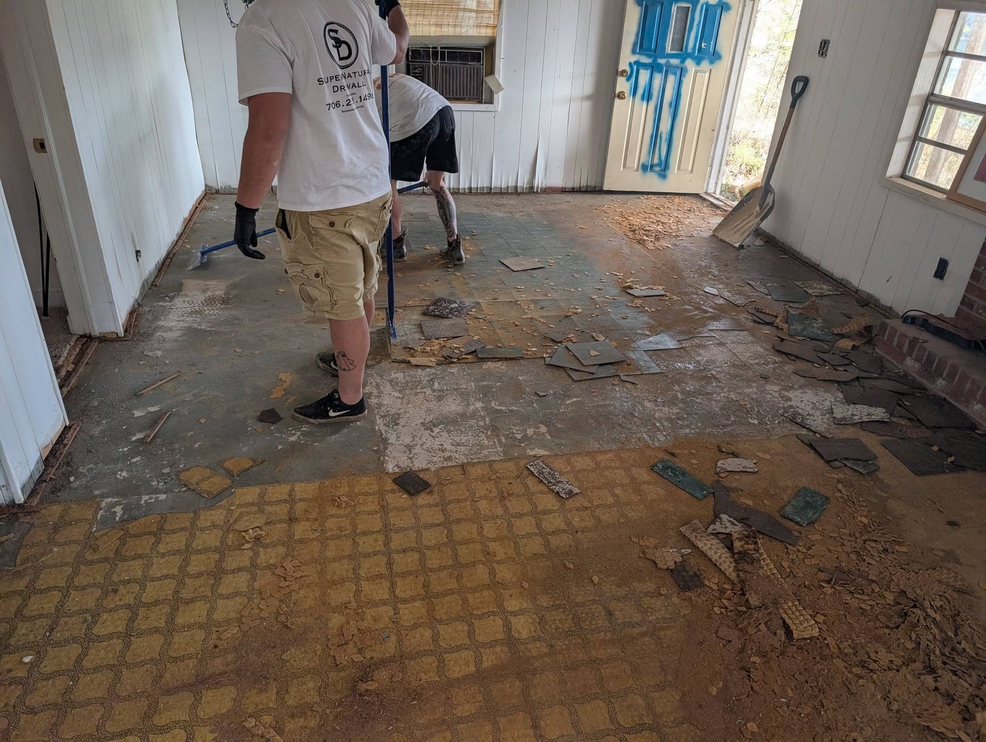 Two people clearing debris from a damaged room with exposed floor and walls
