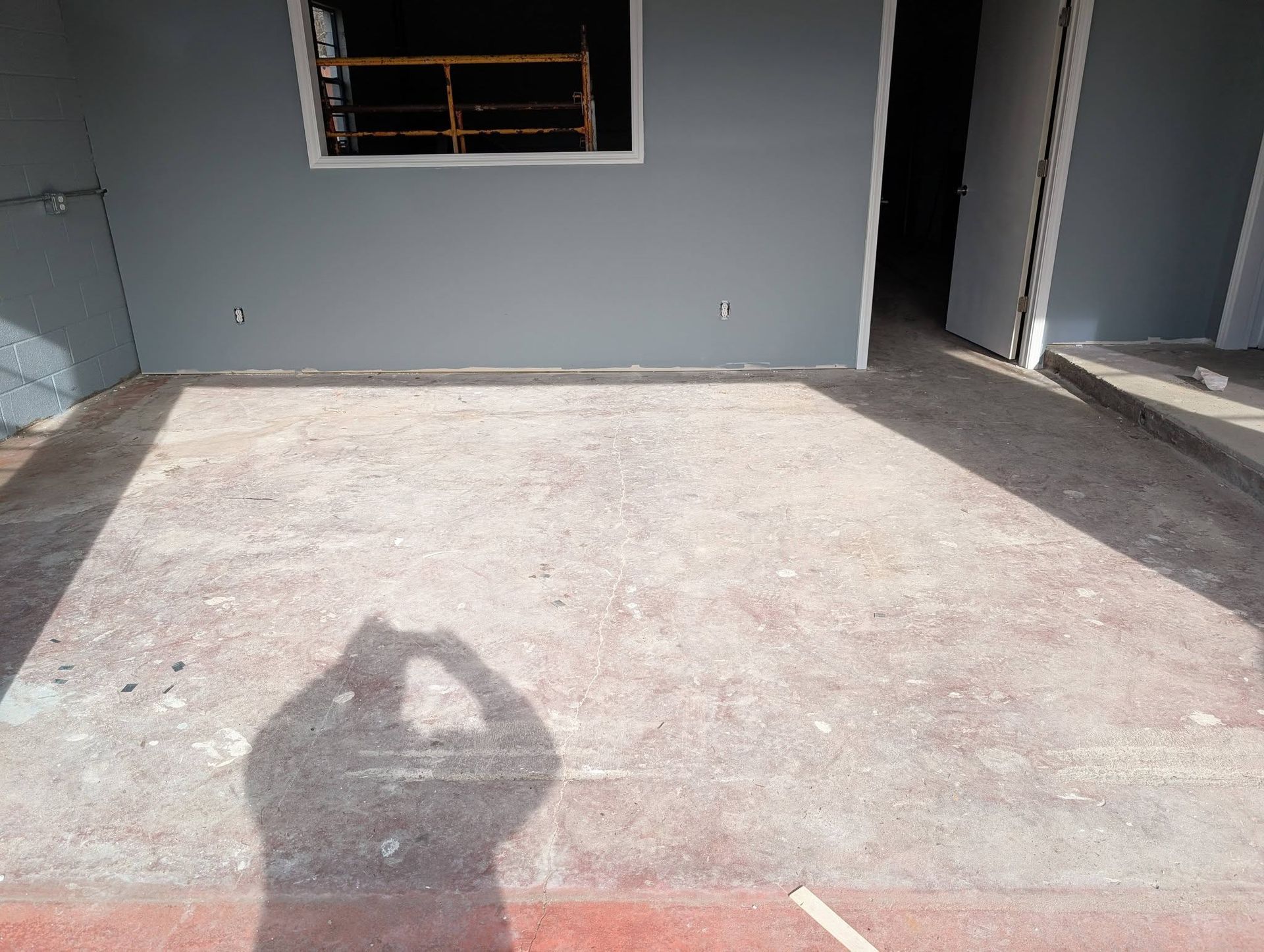 Empty concrete driveway beside gray building with a window and doorway, with a person’s shadow in the foreground