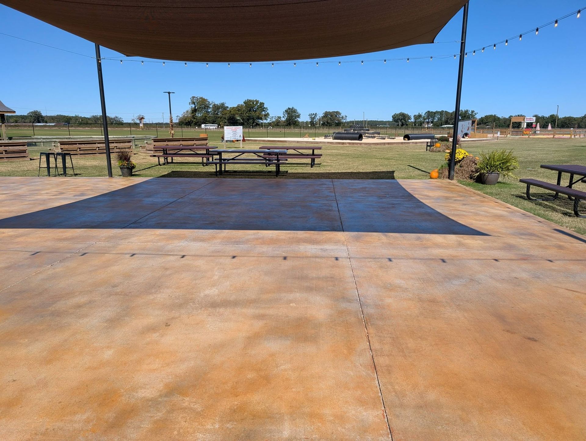 Empty outdoor stage with wooden floor and canopy, facing picnic tables in a park-like field.