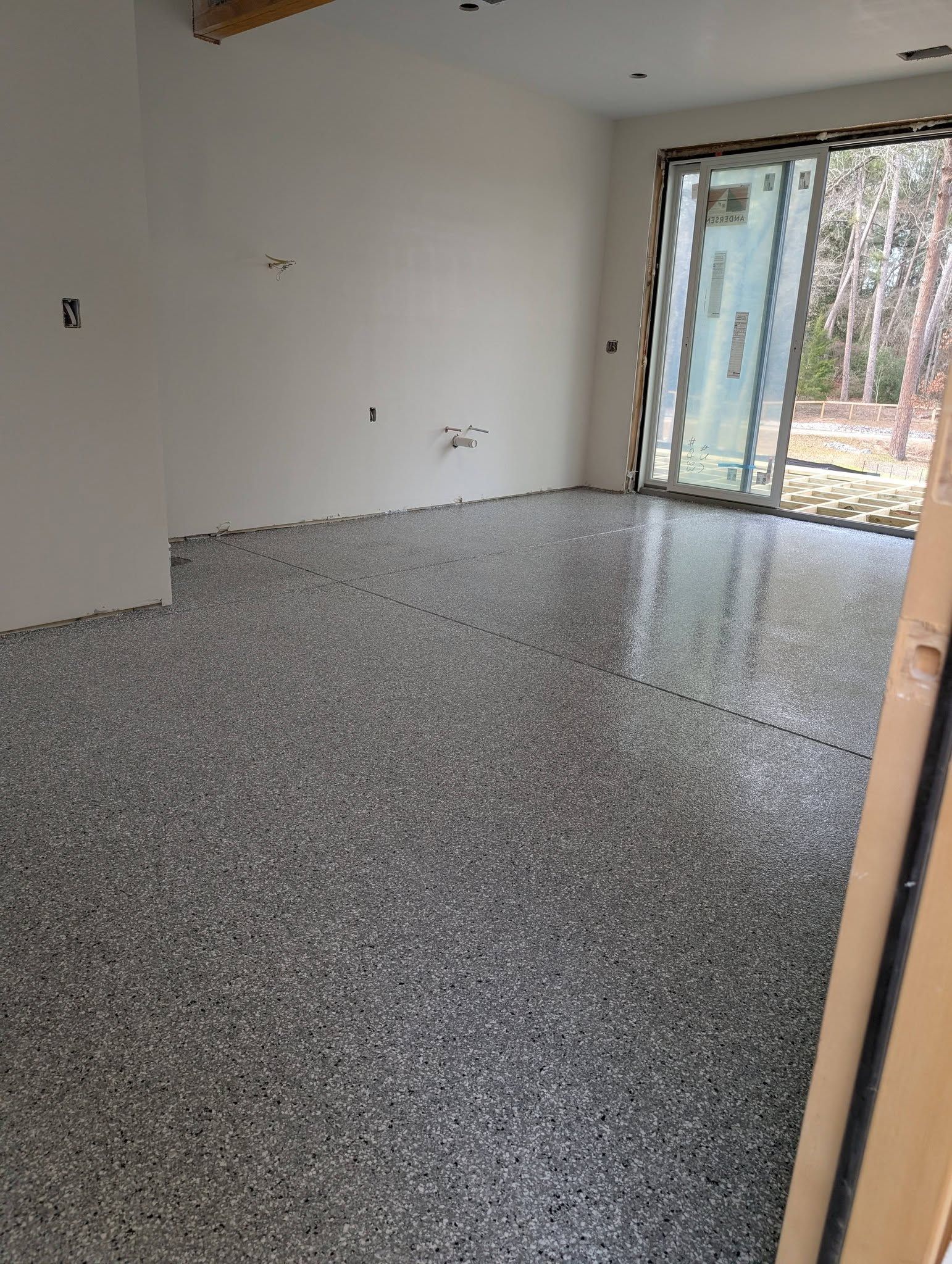 Empty unfurnished room with speckled floor, white walls, and a sliding glass door to a wooded view