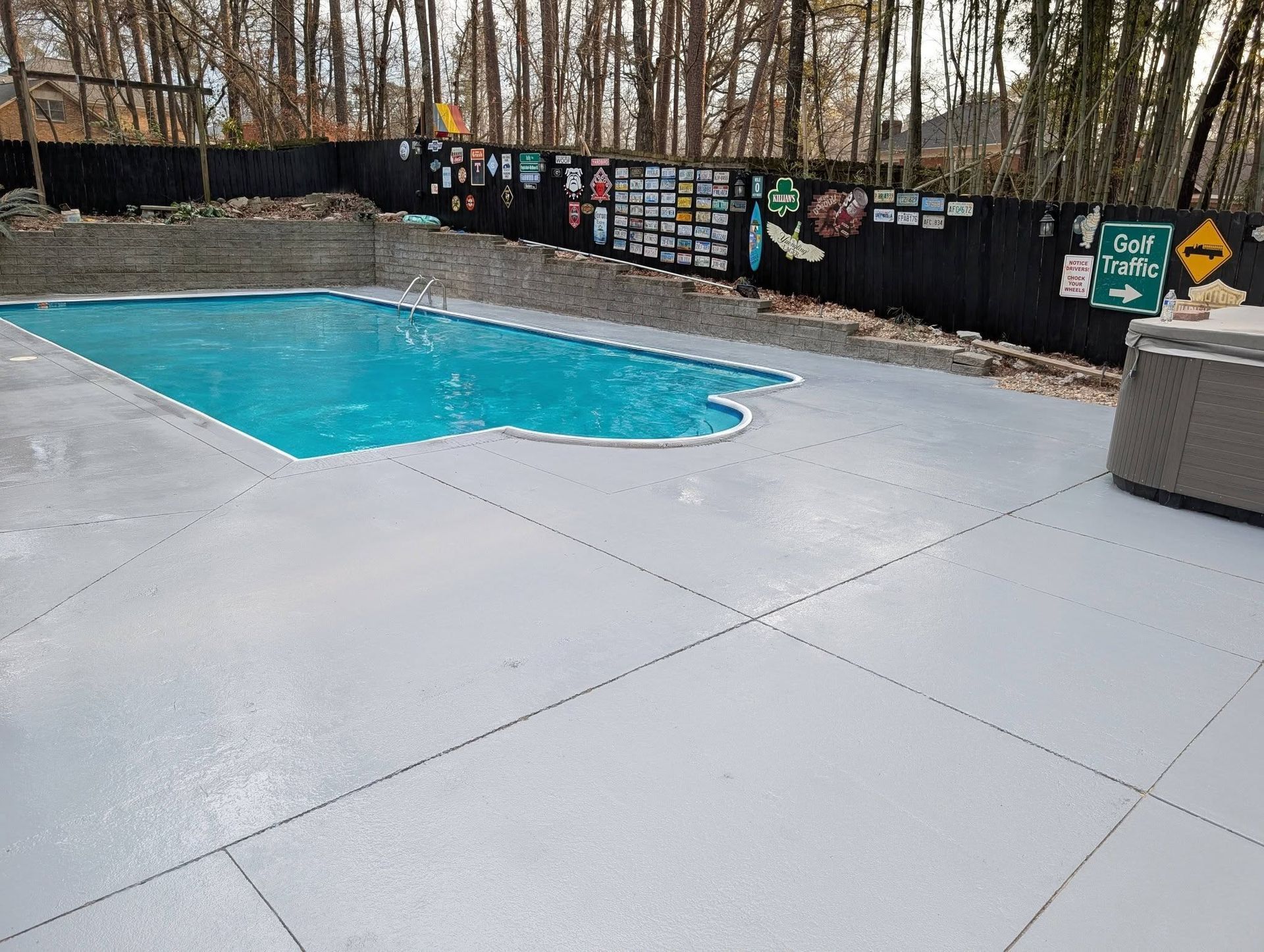 Empty outdoor pool with turquoise water, surrounded by a gray deck and black privacy fence in a wooded area