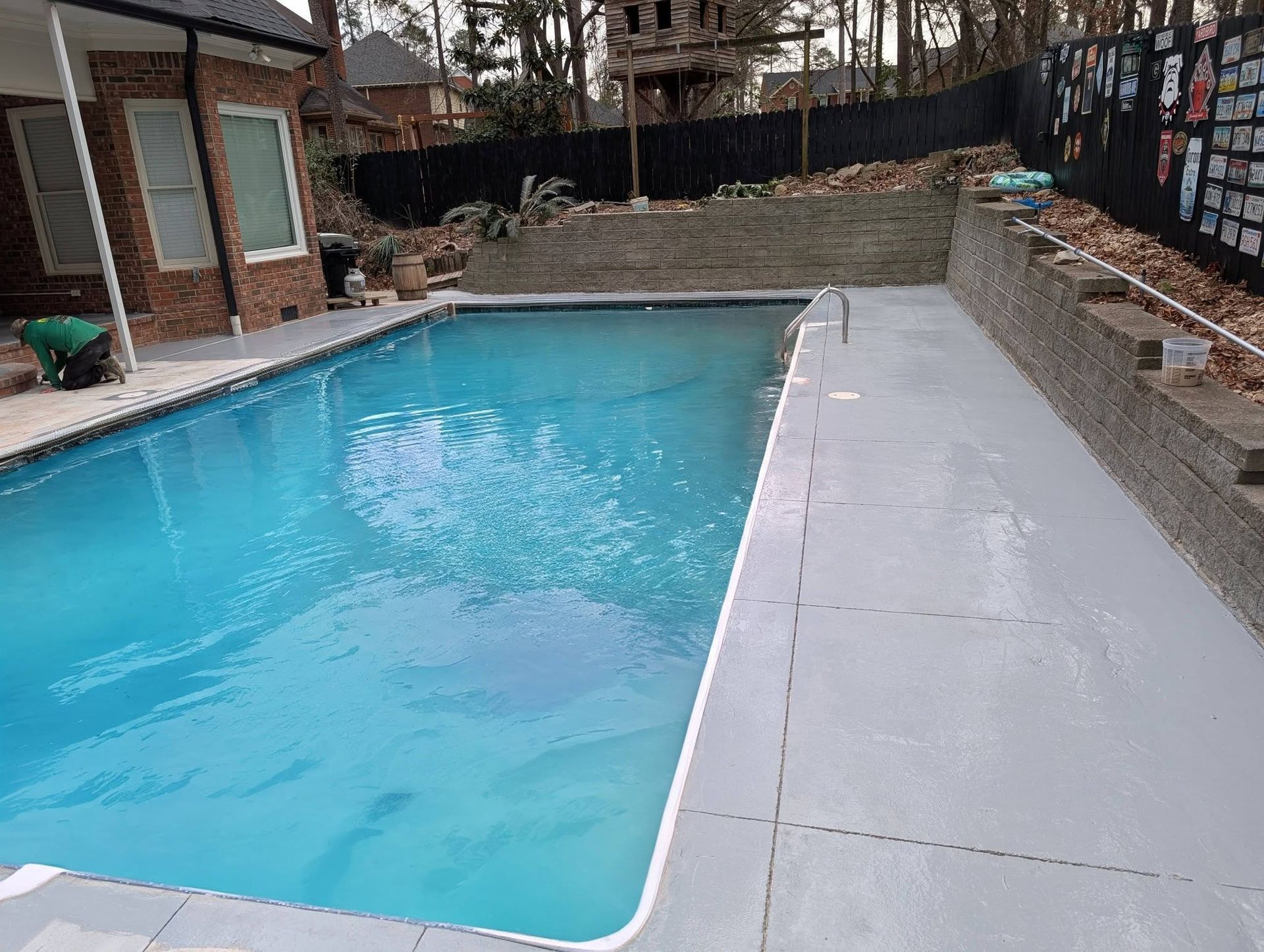 Backyard swimming pool beside a gray concrete deck and brick patio, with a stone retaining wall in back.