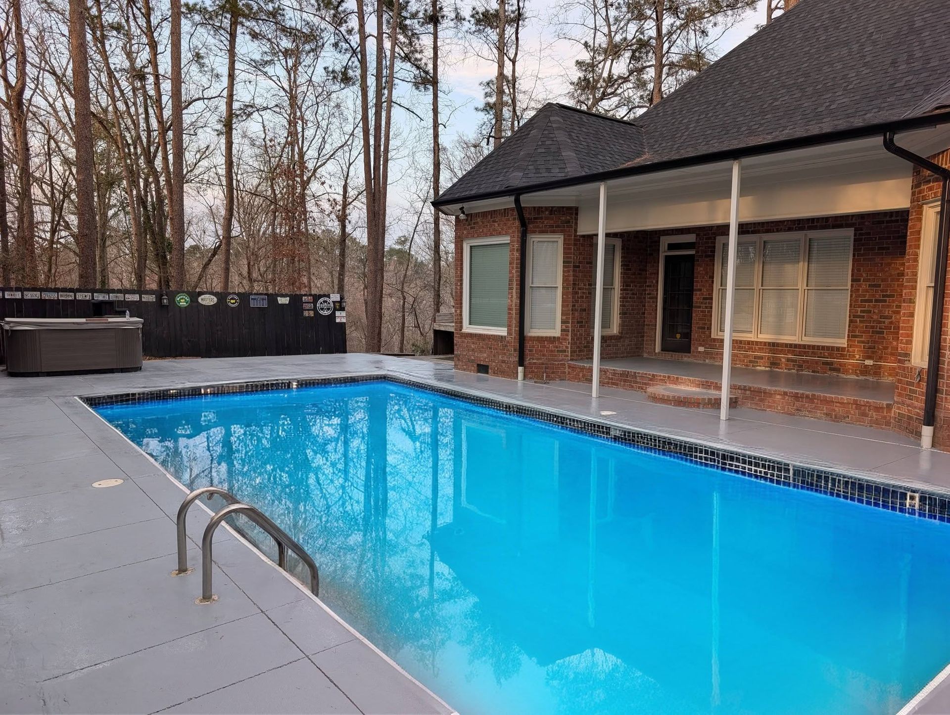 Backyard swimming pool with blue water beside a brick house and wooded fence line