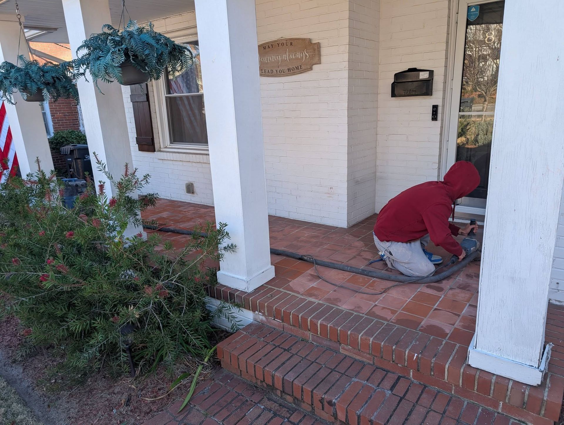 Person in a red hoodie kneeling on a front porch, working near a white column and brick steps.
