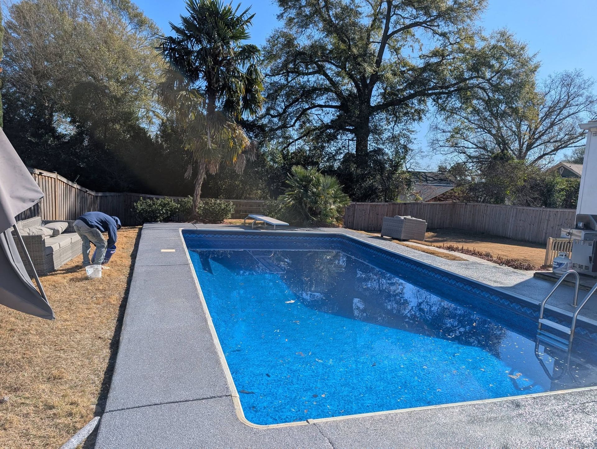 Backyard swimming pool with blue water, stone deck, patio chairs, and trees under a clear sky