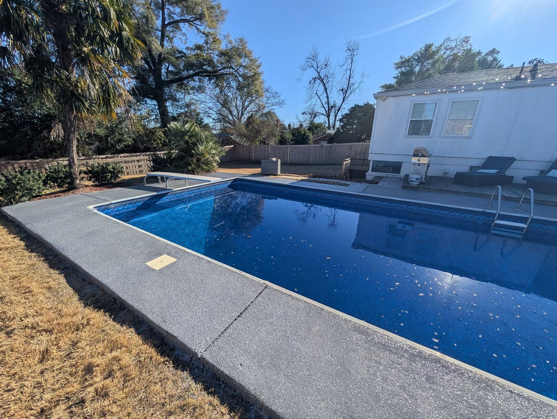 Backyard swimming pool with blue water, concrete deck, patio chairs, and trees under a clear sky