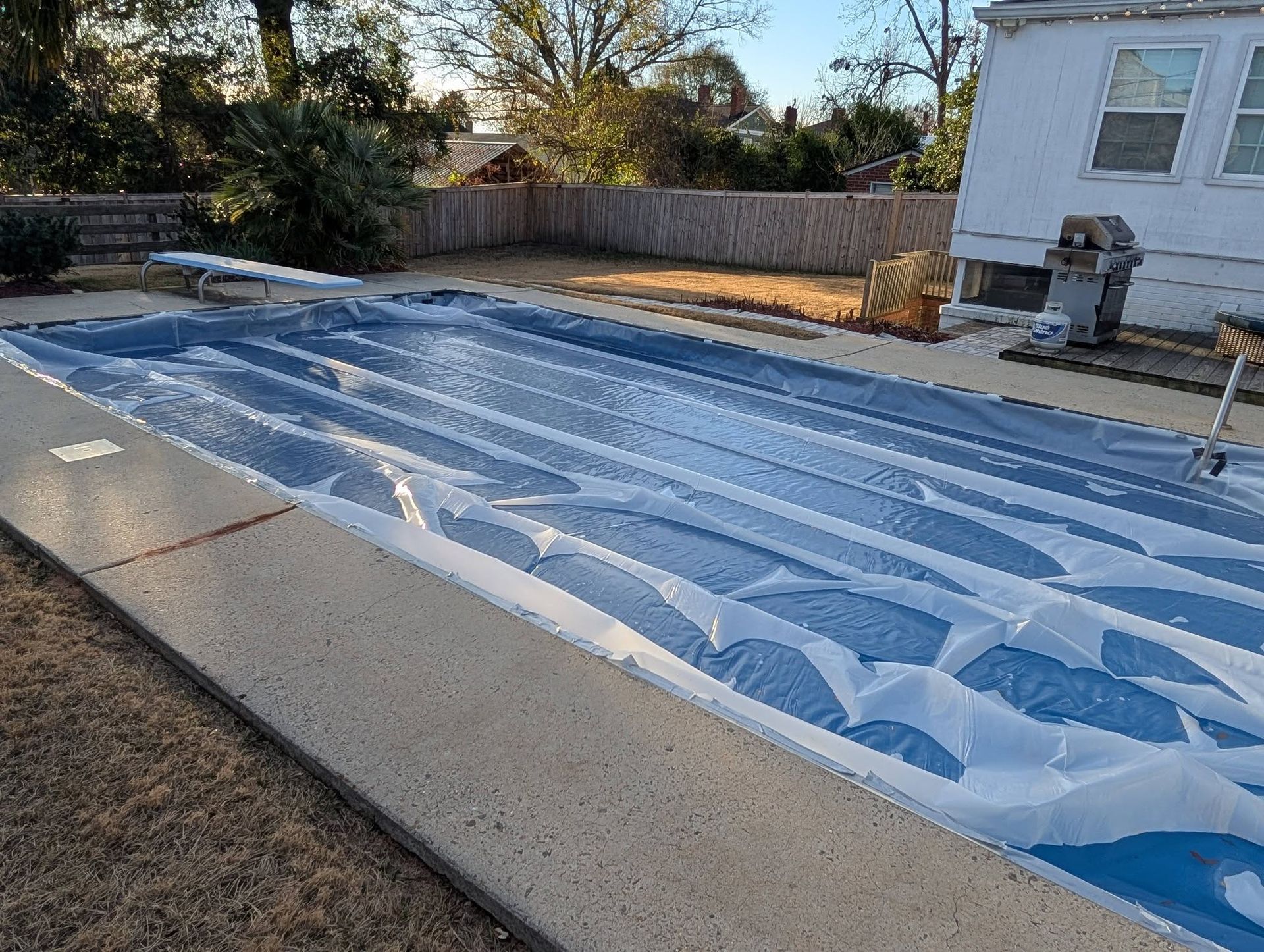 Covered backyard swimming pool with a blue winter tarp beside a house and fence.