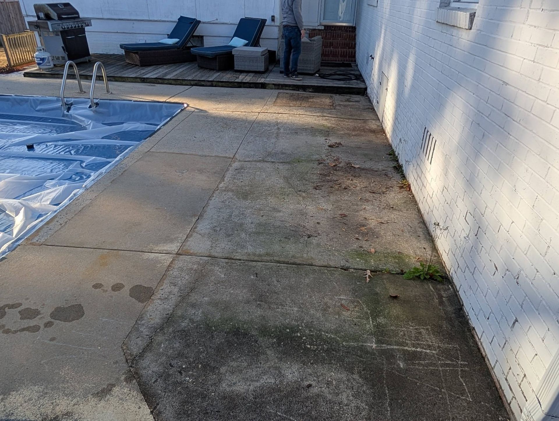 Outdoor patio with concrete walkway, blue-covered pool, lounge chairs, and a brick grill beside a white wall