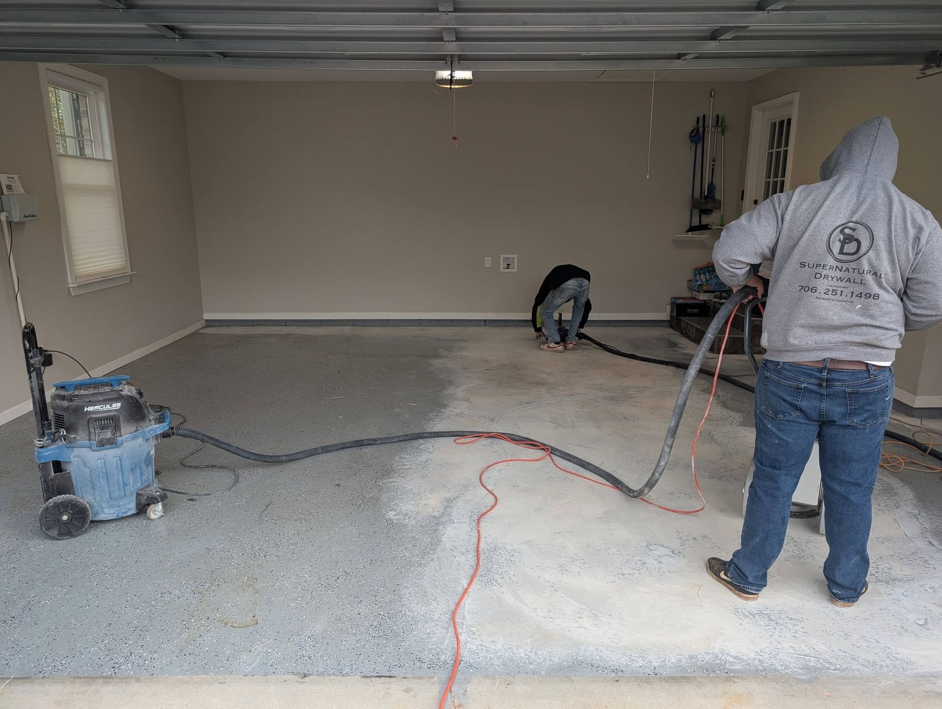 Two workers grind a garage floor, with dust, cords, and tools spread across the concrete.