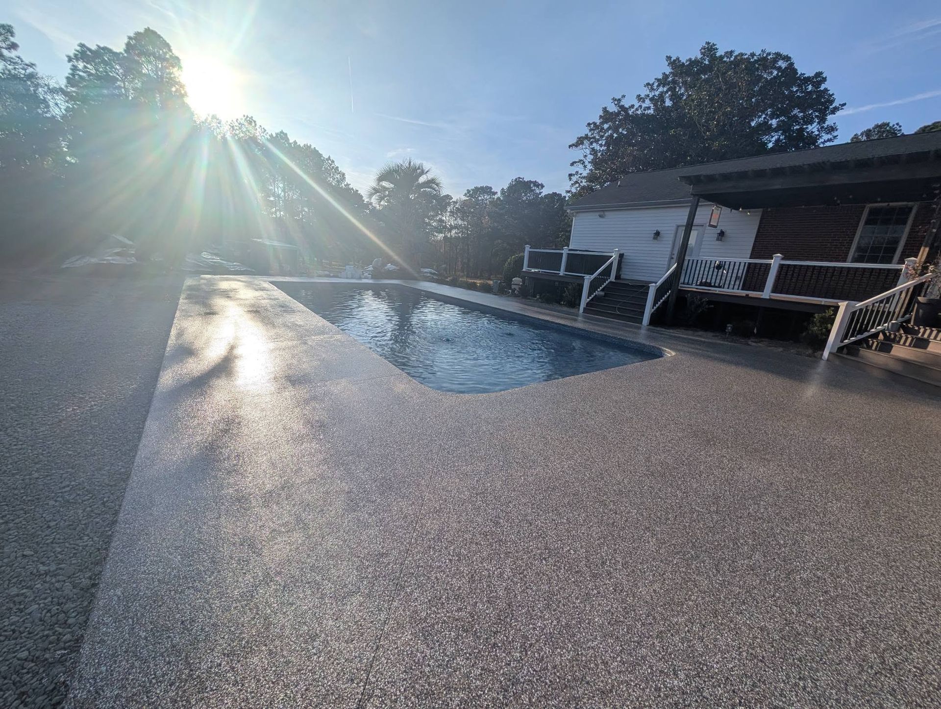 Sunlit backyard pool beside a house with lounge chairs and a wet patio after a rain