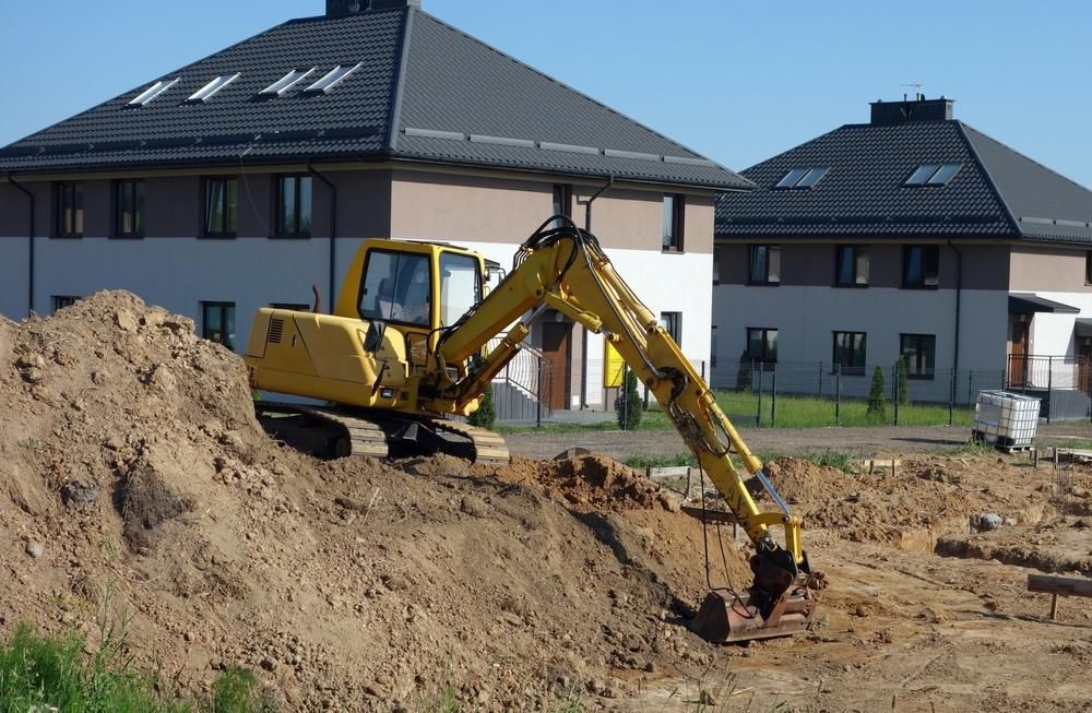 A Yellow Excavator Is Digging A Hole In The Dirt In Front Of Two Houses — Glen Tier Bobcat & Truck Hire In Cardwell, QLD