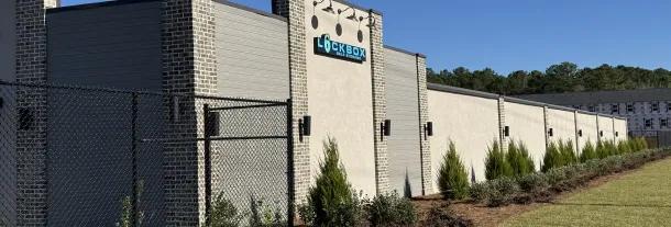 A light-colored building with a logo, featuring a row of green bushes, with a clear blue sky background.