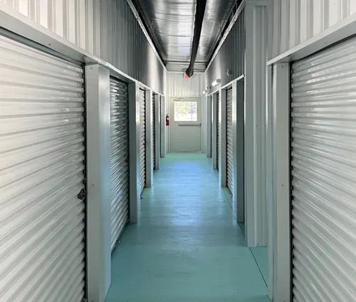 Storage unit hallway with closed white roll-up doors and a blue floor, lit by overhead lights