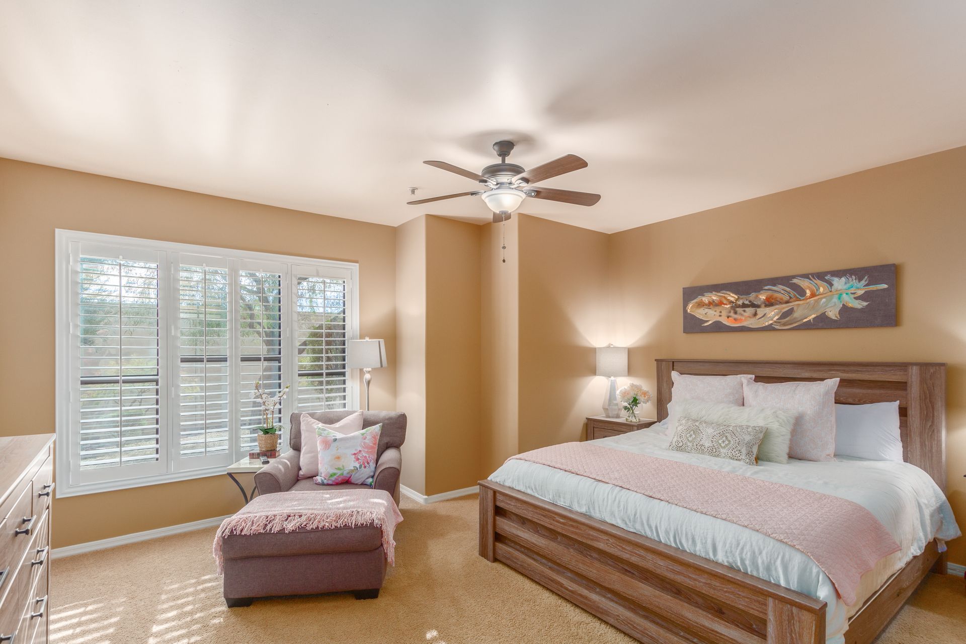 A bedroom with a bed , chair , ottoman and ceiling fan.