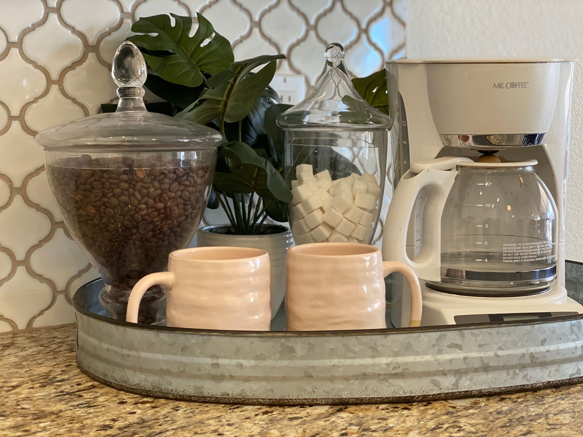 A coffee tray with two cups , a coffee maker , and a jar of coffee beans on a counter.
