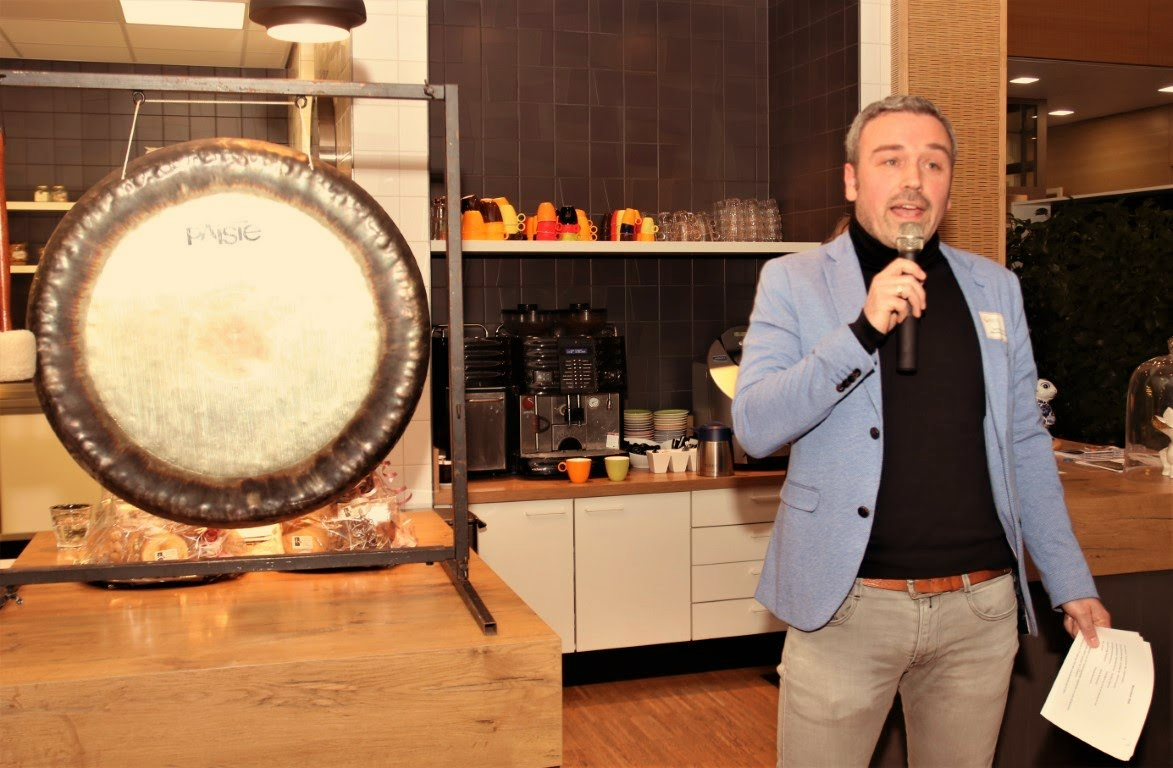 Een man spreekt in een microfoon naast een grote gong in een binnenruimte.