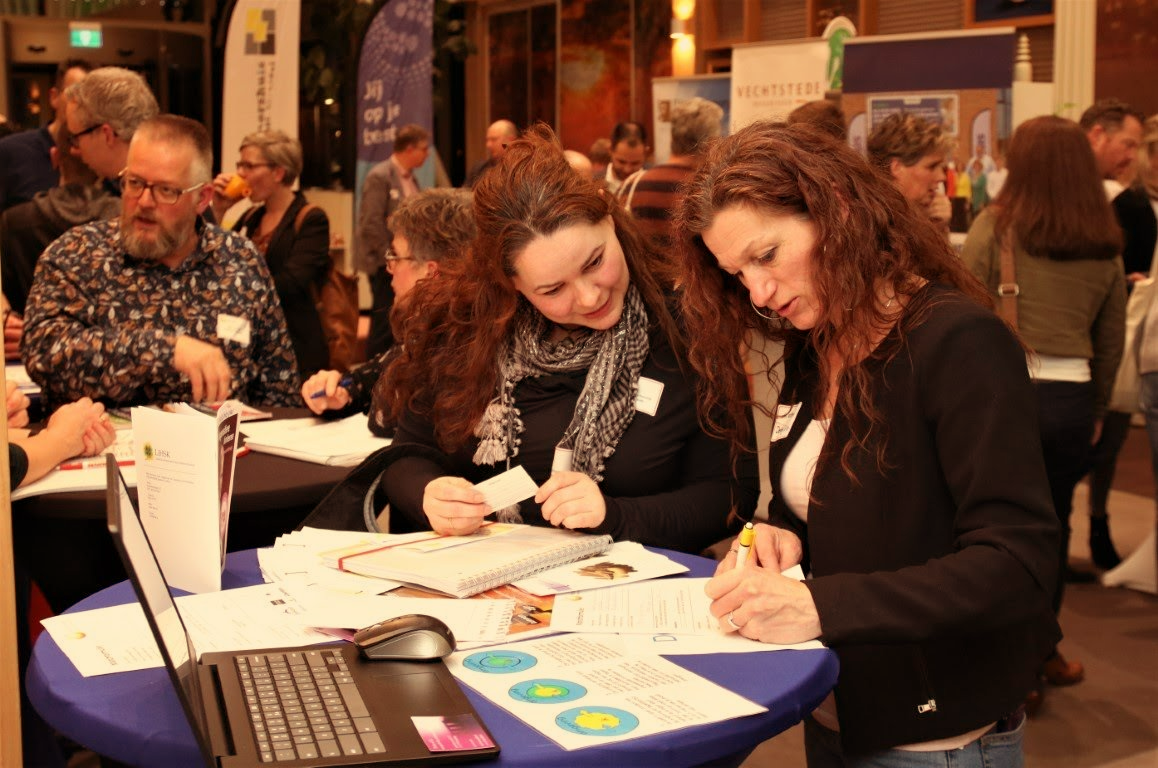 Mensen op een evenement, twee vrouwen die documenten op een tafel bekijken met een laptop, en vele anderen die op de achtergrond rondlopen.