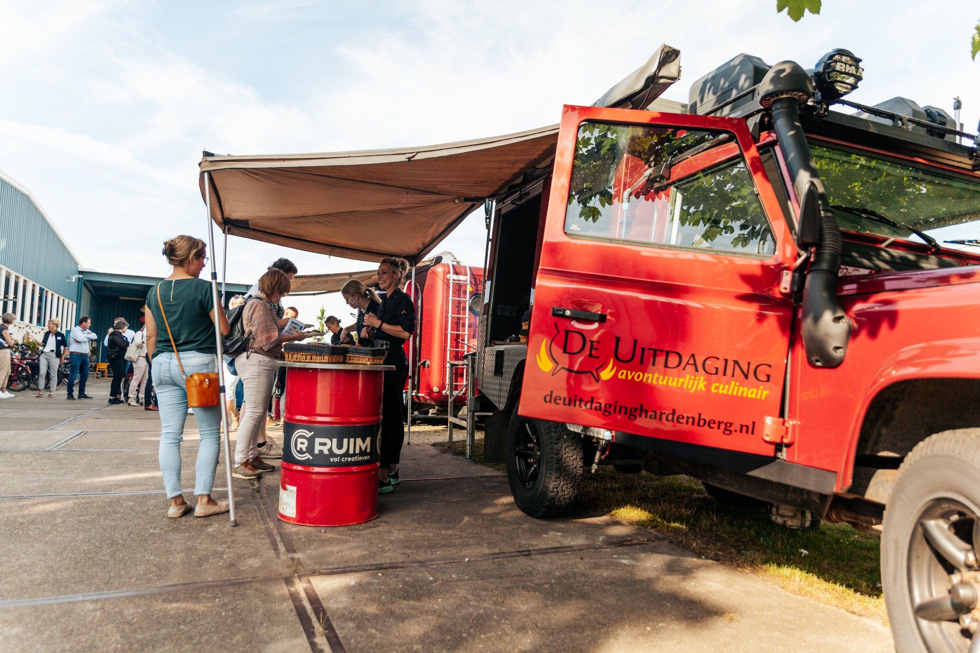 Een rode terreinwagen met luifel serveert eten; mensen verzamelen zich eromheen. Buiten, een zonnige dag.