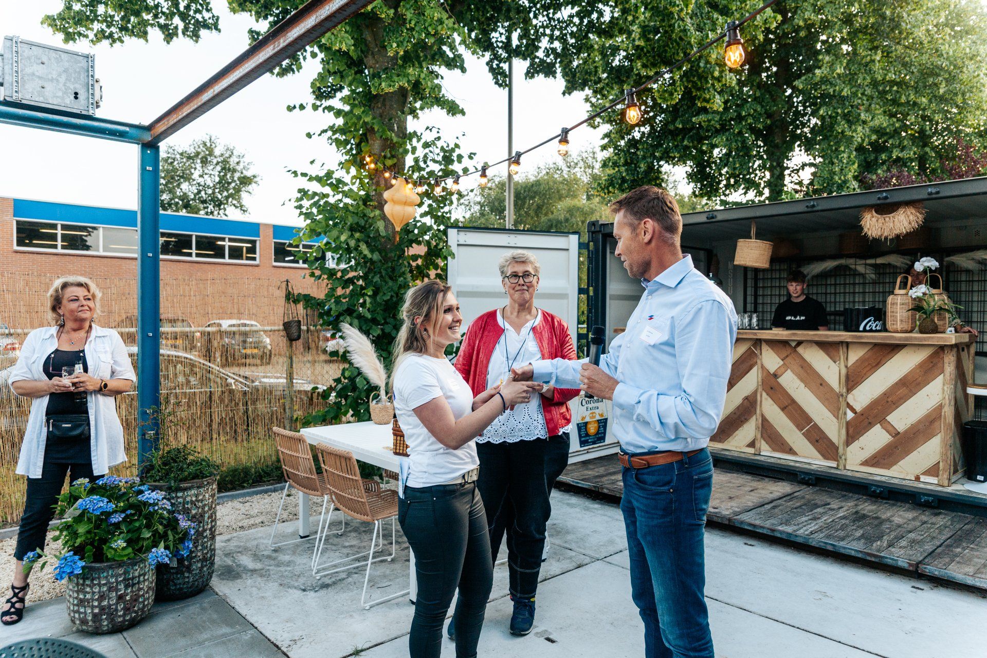 Mensen verzamelen zich in de openlucht bij een bar in een zeecontainer. Een man geeft een vrouw een drankje. Anderen kijken toe.