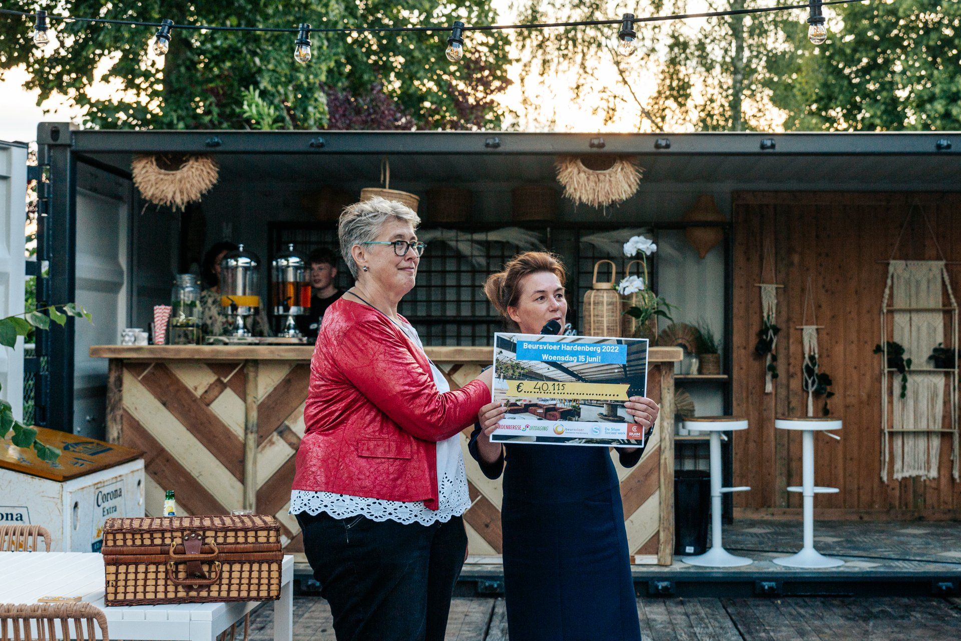 Twee vrouwen presenteren een poster buiten, voor een bar gemaakt van een container.