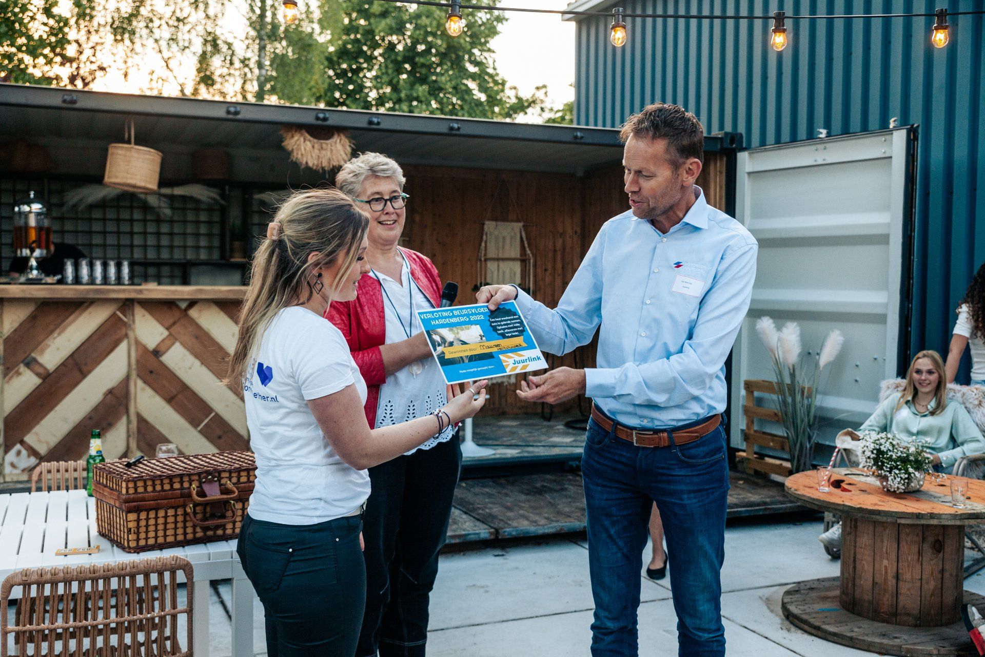 Een man overhandigt een boekje aan een jonge vrouw, terwijl een andere vrouw toekijkt. Buiten, tijdens een evenement.
