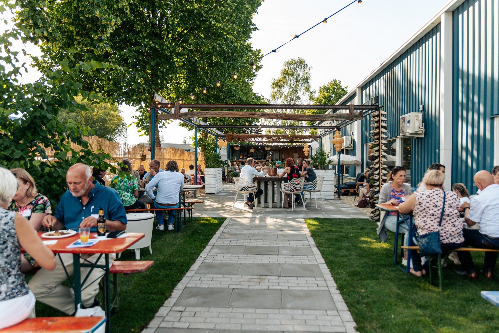 Bijeenkomst in de buitenlucht aan tafels met eten en drinken; grasveld, blauw gebouw op de achtergrond.