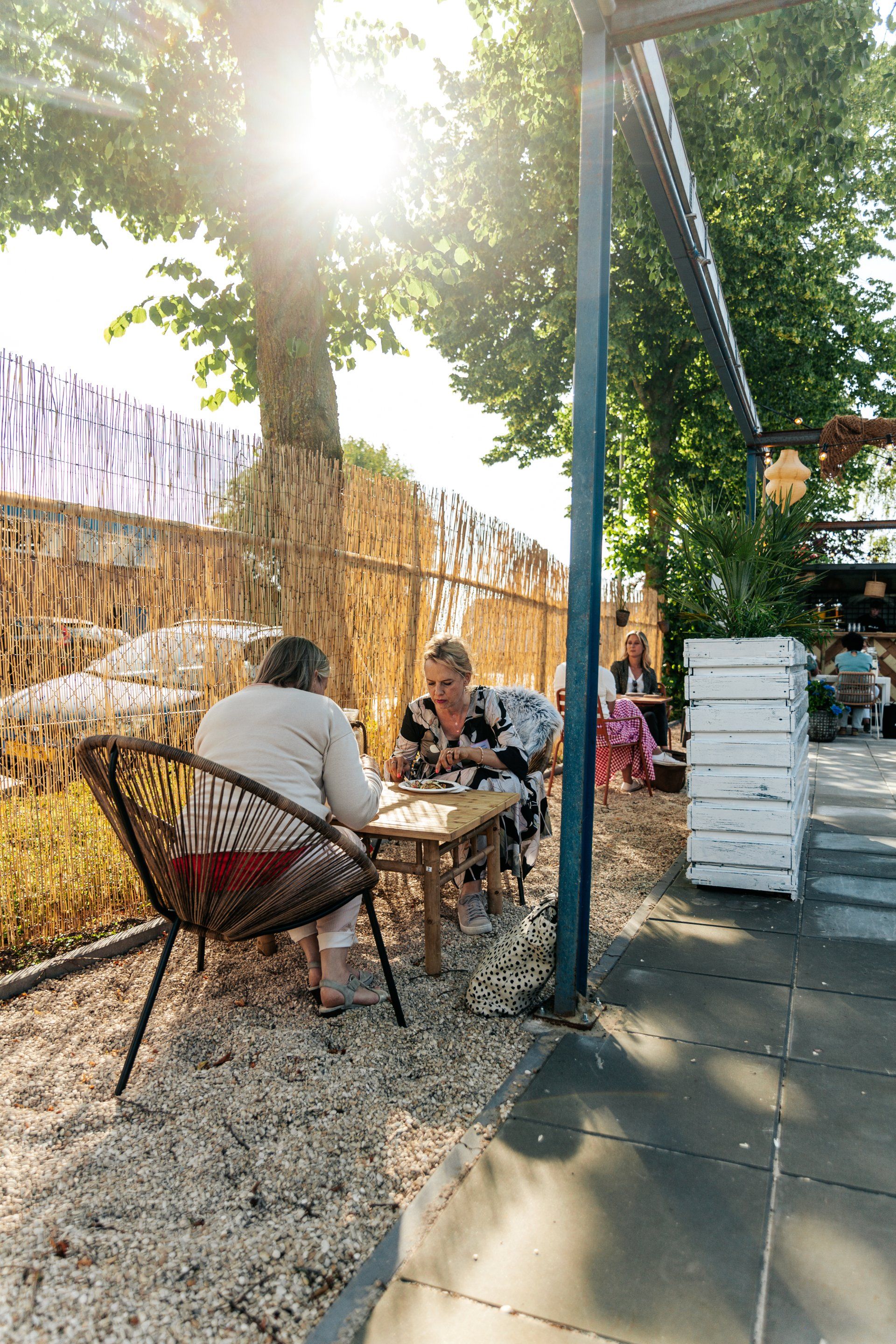Twee mensen zitten buiten aan een tafel, met bomen en zonlicht boven hen, op een grindondergrond.